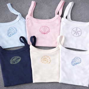 May include: Six tank tops in pastel shades of blue, pink, and white, plus navy and cream. Each top is embroidered with a different seashell design, including a scallop shell and an oyster. The tops have thin straps.