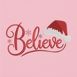 May include: A red embroidered design on a pink background. The word "Believe" is written in a cursive font, with snowflakes and a Santa hat. The hat is red with a white trim and a white pom-pom.
