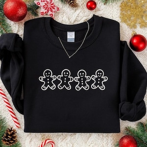 May include: A black sweatshirt featuring a white outline of four gingerbread men. The sweatshirt has a crew neck and long sleeves. The design is centered on the front of the garment. The background includes Christmas ornaments and decorations.