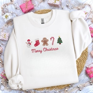 May include: A white sweatshirt with embroidered Christmas-themed designs, including a snowman, stocking, gingerbread man, candy cane, and Christmas tree. The words "Merry Christmas" are embroidered in red below the designs. The sweatshirt has long sleeves.