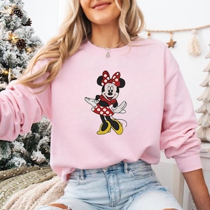 May include: A light pink sweatshirt featuring a Minnie Mouse graphic. The graphic shows Minnie Mouse in a red polka dot dress, red bow, and yellow shoes. The sweatshirt is a long-sleeved crew neck.