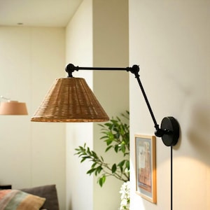 Peut inclure: Une lampe murale avec un abat-jour conique tissé. La lampe est dotée d'un bras réglable noir et d'une base circulaire. L'abat-jour est de couleur marron clair et la lampe est fixée sur un mur blanc.