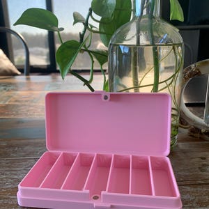May include: A light pink plastic pill organiser with a hinged lid and seven individual compartments. Each compartment is embossed with a single letter representing the days of the week, starting with S for Sunday and ending with S for Saturday.