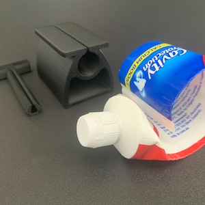 May include: A black plastic toothpaste tube squeezer tool consisting of a T-shaped key and a slotted base, positioned next to a partially used tube of Colgate Cavity Protection Calcium Boost toothpaste with a white cap and blue and red branding.
