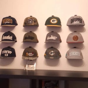 May include: A collection of eighteen baseball caps arranged in three rows on a wall. The caps feature various colours including black, navy, grey, green, and red, with diverse embroidered logos, text such as DROPPED, HWDP, and ZUCKER, and patterns.