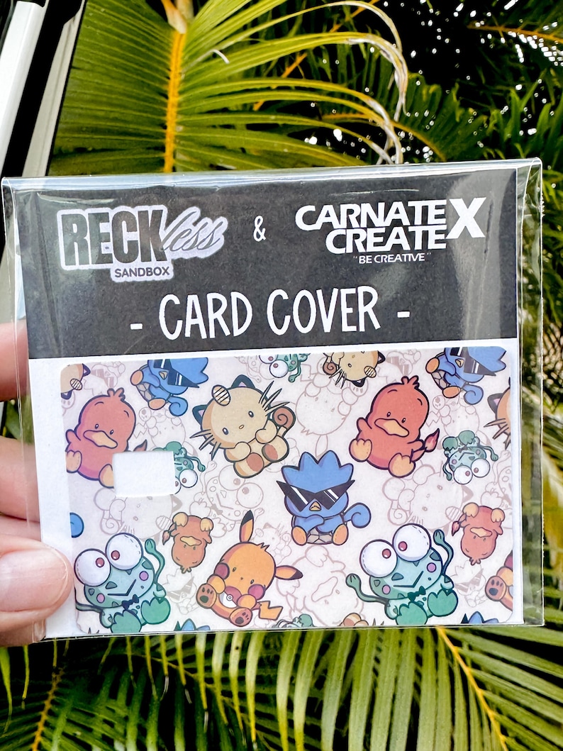 May include: A clear plastic card cover with a vibrant pattern of cartoon characters. The card cover has the text "RECKLESS & CARNATE X CREATE - CARD COVER -" printed on it. The card cover is held in a person's hand.