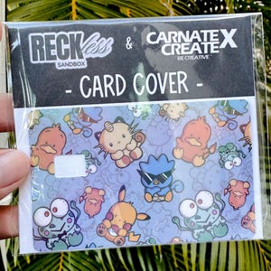 May include: A card cover in a plastic sleeve, patterned with cartoon characters. The card cover has a light blue background with various cartoon characters. The text "RECKLESS & CARNATE X CREATE - CARD COVER - SANDBOX" is printed on the top.