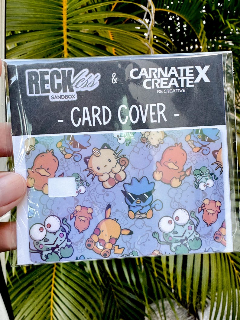 May include: A card cover in a plastic sleeve, patterned with cartoon characters. The card cover has a light blue background with various cartoon characters. The text "RECKLESS & CARNATE X CREATE - CARD COVER - SANDBOX" is printed on the top.