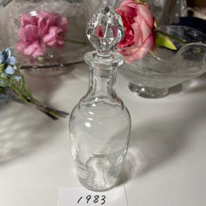 May include: A clear glass perfume bottle with a faceted crystal stopper. The bottle has a slender, elegant shape. A white label with the number "1983" is visible.