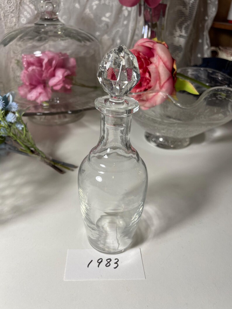 May include: A clear glass perfume bottle with a faceted crystal stopper. The bottle has a slender, elegant shape. A white label with the number "1983" is visible.