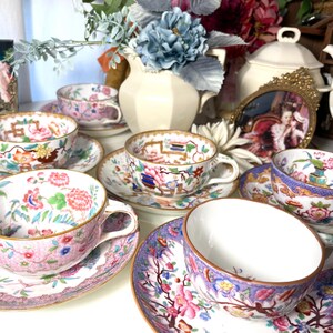 May include: A set of vintage teacups and saucers featuring floral designs in pink, blue, and gold. The teacups are displayed on a white surface, with a blurred backdrop of flowers and a decorative frame.