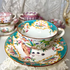 May include: A porcelain teacup and saucer set featuring a white base with a vibrant teal border, gold scrollwork, and multi-colored floral and botanical motifs. The cup has a curved handle and a brown rim, with additional teacups visible in the background.