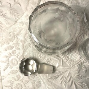 May include: A clear, faceted glass jar with a matching stopper. The jar has a geometric design and is sitting on a white, textured surface. The stopper is round with a faceted design.