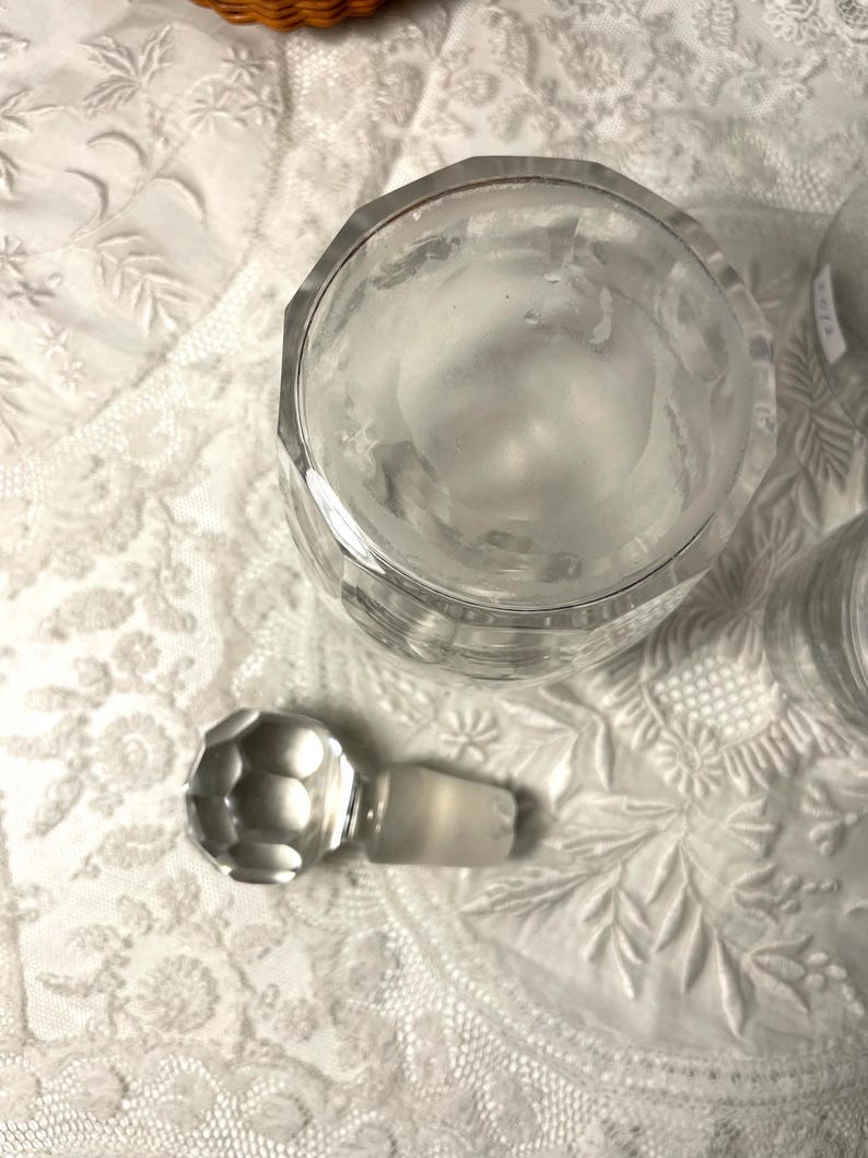May include: A clear, faceted glass jar with a matching stopper. The jar has a geometric design and is sitting on a white, textured surface. The stopper is round with a faceted design.