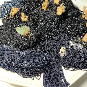 Puede incluir: Una colección de hebras de cuentas en varios tonos de azul y negro, dispuestas sobre un plato blanco con borde festoneado. Las cuentas parecen pequeñas y ensartadas, con algunas hebras con borlas.