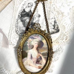 Peut inclure: Un portrait ovale ancien dans un cadre doré, suspendu par une chaîne. Le portrait représente une femme dans une robe et un chapeau pastel. Le cadre est orné de détails floraux. Une estampe religieuse est visible en arrière-plan.