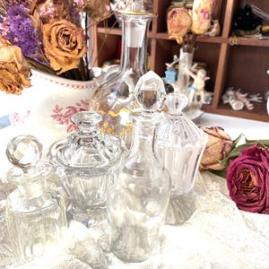 May include: A collection of vintage glass perfume bottles and jars with crystal stoppers. The bottles vary in shape and size, with intricate detailing. Dried roses and other flowers are in the background, adding a touch of antique charm. The bottles are displayed on a white lace cloth.