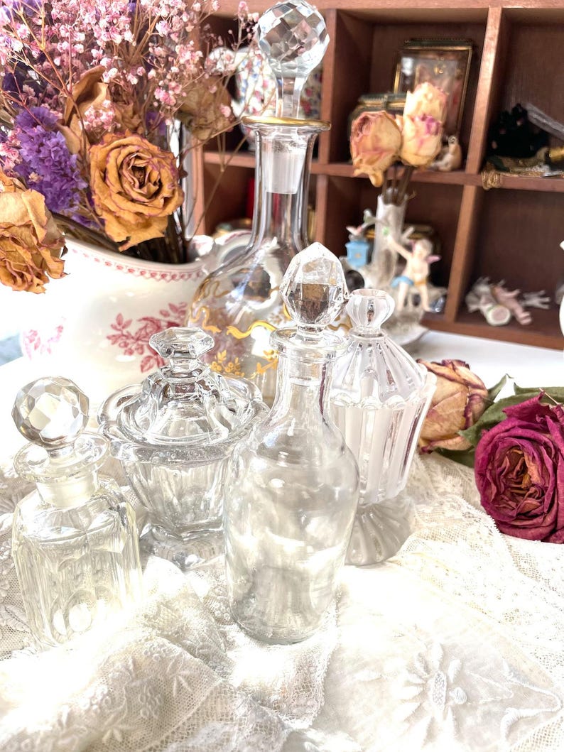 May include: A collection of vintage glass perfume bottles and jars with crystal stoppers. The bottles vary in shape and size, with intricate detailing. Dried roses and other flowers are in the background, adding a touch of antique charm. The bottles are displayed on a white lace cloth.