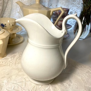 Op de afbeelding: Een witte keramische kan met een gebogen tuit en een lusvormige handgreep. De kan heeft een bolvormig lichaam en een decoratieve band rond het midden. Andere vintage theeserviesstukken zijn zichtbaar op de achtergrond, waaronder een theepot en kopjes.