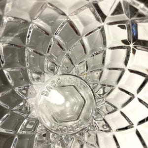May include: Close-up of a clear glass object with a diamond-cut pattern. The center features a circular emblem with the text "BACCARAT DEPOSE" in a hexagon shape. The glass reflects light, creating a sparkling effect.