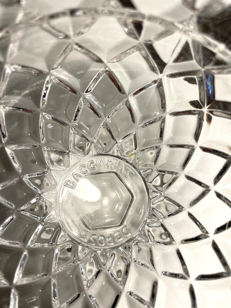 May include: Close-up of a clear glass object with a diamond-cut pattern. The center features a circular emblem with the text "BACCARAT DEPOSE" in a hexagon shape. The glass reflects light, creating a sparkling effect.