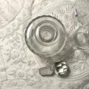 May include: Clear glass bottle with a faceted stopper. The bottle has a wide, circular base and a narrow neck. The stopper is a geometric shape. The bottle and stopper are on a white, patterned surface.