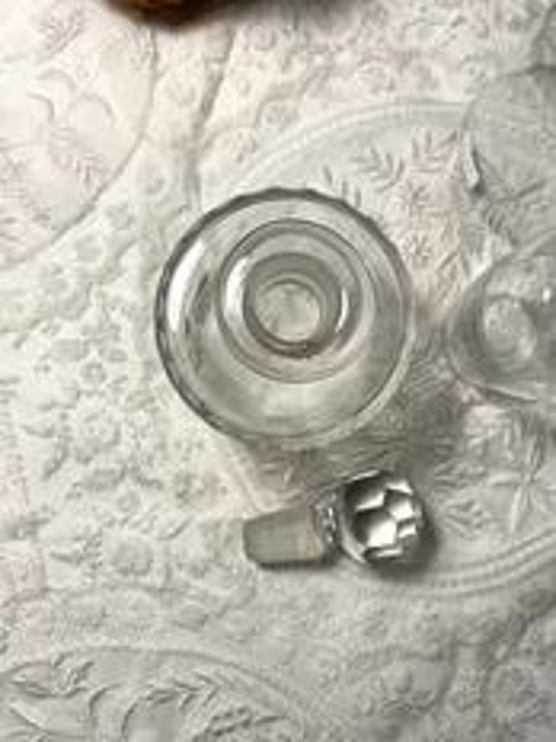 May include: Clear glass bottle with a faceted stopper. The bottle has a wide, circular base and a narrow neck. The stopper is a geometric shape. The bottle and stopper are on a white, patterned surface.