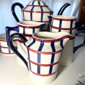 Może przedstawiać: Biały ceramiczny zestaw do herbaty z niebiesko-czerwonym wzorem w paski. Zestaw zawiera czajniczek, dzbanuszek do śmietanki, cukiernicę i filiżanki. Uchwyty i wylewki są granatowe.