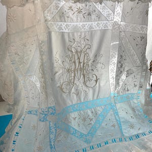 May include: A vintage white and light blue textile with detailed embroidery and lace. It features a central monogram, floral patterns, and geometric designs. The edges are trimmed with delicate lace and a light blue border.