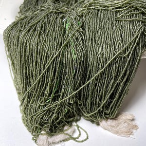 Puede incluir: Primer plano de cuentas de semillas verde oliva, ensartadas juntas, formando hebras largas. Las cuentas se muestran en un plato blanco, con los extremos de las hebras atados con hilo blanquecino. Las cuentas tienen un ligero brillo.