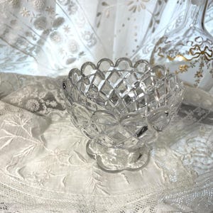 May include: A clear, cut-glass bowl with a scalloped rim and diamond pattern. The bowl sits on a small, round base. The background includes a decorative glass bottle and white lace fabric.