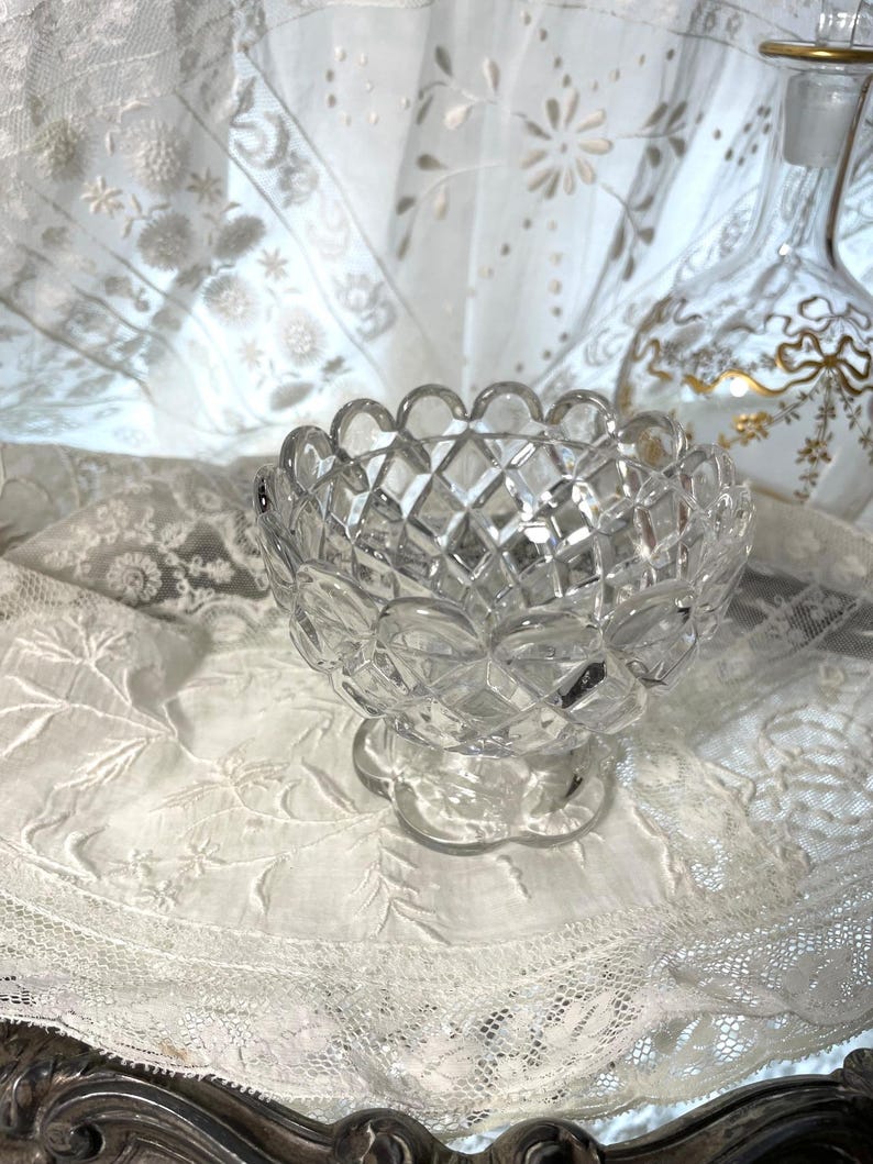 May include: A clear, cut-glass bowl with a scalloped rim and diamond pattern. The bowl sits on a small, round base. The background includes a decorative glass bottle and white lace fabric.