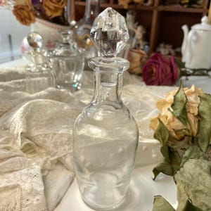 May include: A clear glass perfume bottle with a faceted crystal stopper. The bottle is cylindrical with a narrow neck. Dried roses and other vintage perfume bottles are in the background. The overall theme is vintage and antique.