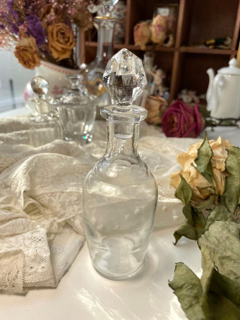 May include: A clear glass perfume bottle with a faceted crystal stopper. The bottle is cylindrical with a narrow neck. Dried roses and other vintage perfume bottles are in the background. The overall theme is vintage and antique.