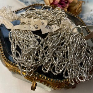 Puede incluir: Hebras de cuentas plateadas se exhiben en una caja vintage ornamentada con bordes dorados. Las cuentas son pequeñas y agrupadas, con etiquetas adjuntas. Una rosa seca y un plato decorativo están en el fondo, añadiendo estética antigua.