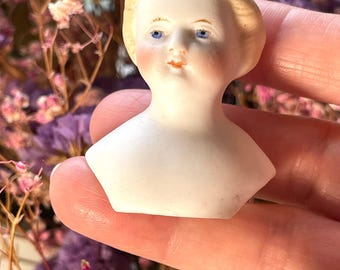May include: A porcelain doll head with a pale complexion, blue eyes, and rosy cheeks. The doll's hair is a light blonde color, styled in a simple, classic fashion. The doll head is a vintage item, likely from a larger doll.