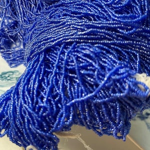 Op de afbeelding: Close-up van een grote hoeveelheid kleine, helderblauwe kralen. De kralen zijn aan elkaar geregen, waardoor een vloeiend, gestructureerd uiterlijk ontstaat. De kralen zijn waarschijnlijk voor het maken van sieraden of knutselprojecten.