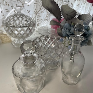 May include: A collection of clear glass decorative items, including a lidded candy dish, a small bowl, and two perfume bottles with stoppers. The items have diamond-cut patterns and are arranged on a white surface.