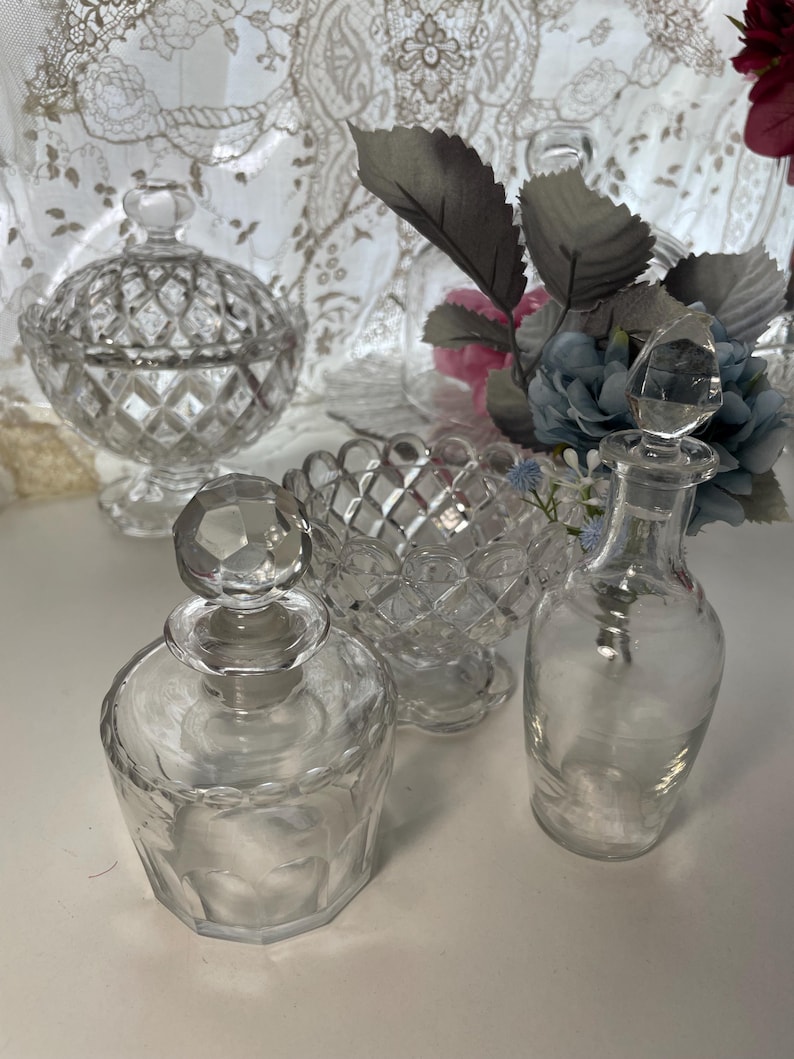 May include: A collection of clear glass decorative items, including a lidded candy dish, a small bowl, and two perfume bottles with stoppers. The items have diamond-cut patterns and are arranged on a white surface.