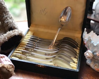 May include: A collection of silver spoons presented in a black box with a cream-coloured lining. One spoon is positioned upright, highlighting the decorative handle. The open box displays the spoon arrangement, indicating a vintage or antique set.