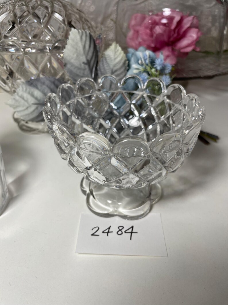 May include: A clear, diamond-cut glass bowl with a scalloped rim and a pedestal base. The bowl has a textured, geometric pattern. A small white tag with the number "2484" is visible.