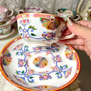 May include: A white porcelain teacup and saucer set featuring a colourful hand-painted pattern of pink flowers, blue scrolling vines, and orange fan-shaped motifs. The rim of the saucer and the cup are accented with a vibrant orange and pink border.