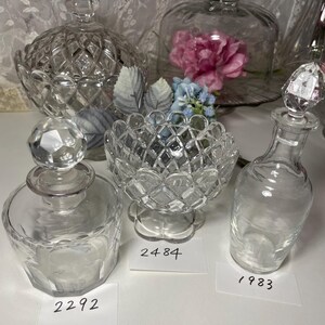 May include: A collection of clear glass decorative items, including a lidded candy dish, a small bowl, and a perfume bottle with a faceted stopper. The items have diamond-cut patterns and are arranged on a white surface.