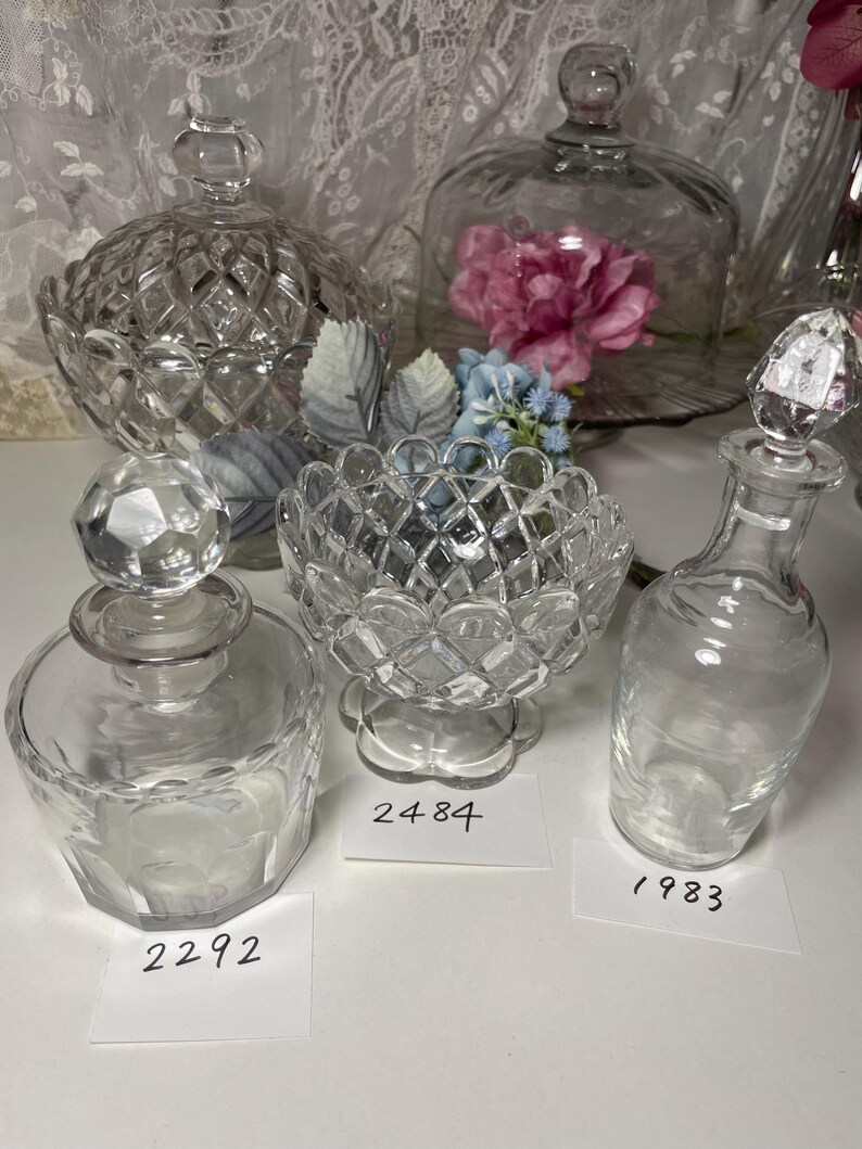 May include: A collection of clear glass decorative items, including a lidded candy dish, a small bowl, and a perfume bottle with a faceted stopper. The items have diamond-cut patterns and are arranged on a white surface.
