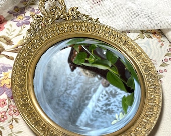 May include: An antique hand mirror with a gold-toned frame and handle. The round mirror is encircled by intricate floral and scroll detailing. The handle features a decorative, fluted design. The mirror reflects a green plant and other objects.