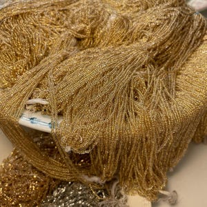 Könnte beinhalten: Nahaufnahme eines Haufens goldener Perlenstränge, wahrscheinlich zum Basteln oder Verzieren. Die Perlen sind klein und reflektierend und erzeugen einen schimmernden Effekt. Eine kleine Menge silberner Perlenstränge ist ebenfalls sichtbar.