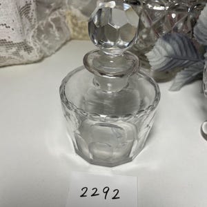 May include: A clear glass perfume bottle with a faceted, geometric design. The bottle has a round base and a faceted stopper. The number "2292" is printed on a white label in the foreground. The bottle is likely vintage.