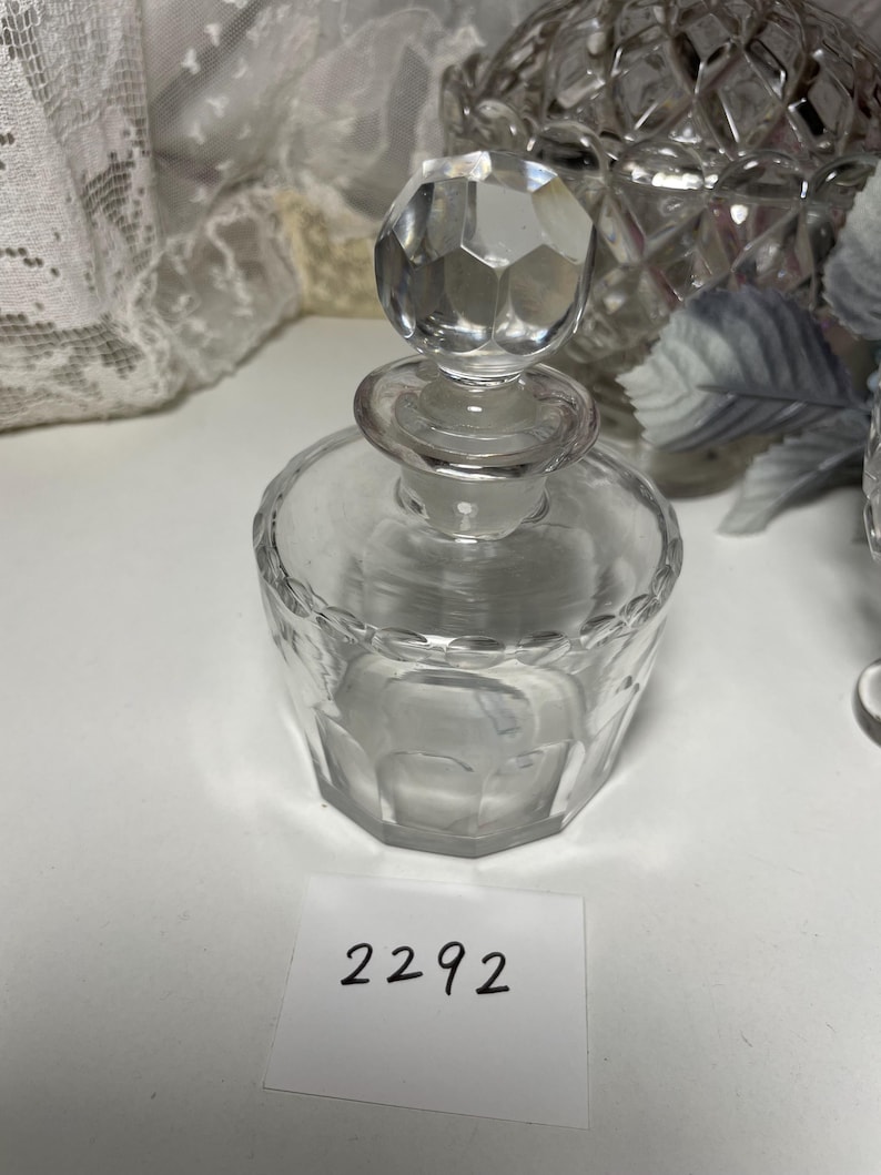 May include: A clear glass perfume bottle with a faceted, geometric design. The bottle has a round base and a faceted stopper. The number "2292" is printed on a white label in the foreground. The bottle is likely vintage.