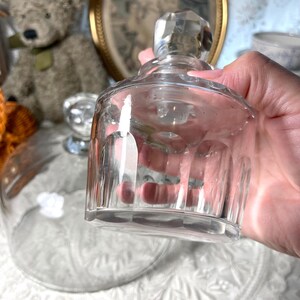 May include: A clear glass bottle with a faceted stopper is held in a hand. The bottle has a cylindrical body with vertical ridges. A glass cloche is in the background, along with a teddy bear and a framed picture.