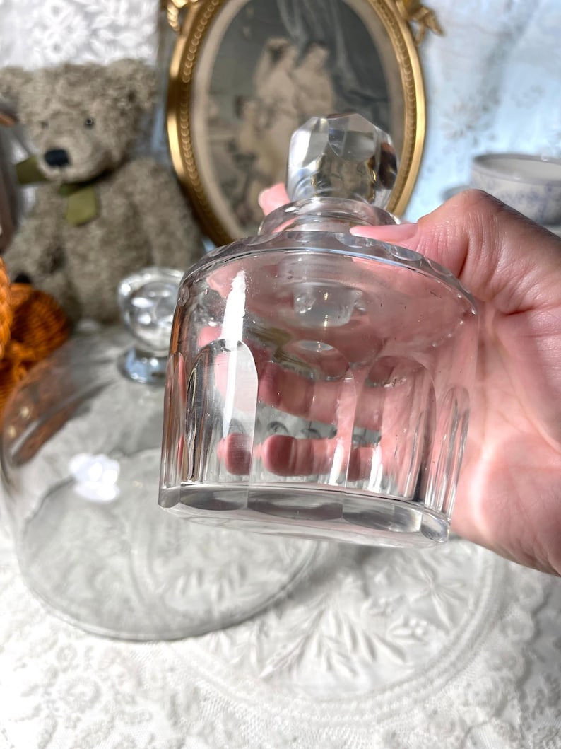 May include: A clear glass bottle with a faceted stopper is held in a hand. The bottle has a cylindrical body with vertical ridges. A glass cloche is in the background, along with a teddy bear and a framed picture.